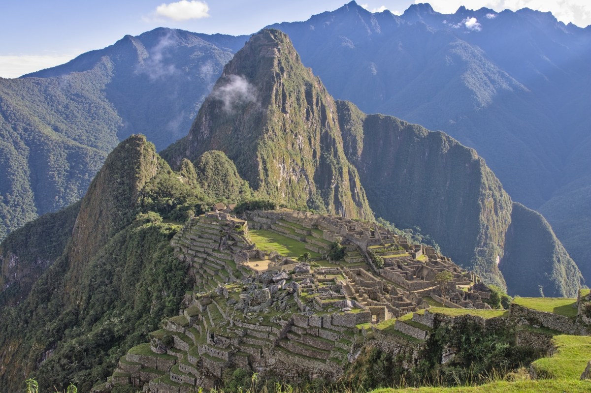 Ancient stone ruins on a mountain ridge. The image shows Machu Picchu, the famous Inca citadel in Peru. The ruins consist of terraced stone walls, stairways, and buildings, all built on a steep, green mountainside. In the background, there are dramatic, lush green mountains rising sharply, with a prominent peak (Huayna Picchu) towering behind the site. The sky is mostly clear with a few clouds, and the overall scene is vibrant and majestic, highlighting the remote and elevated location of this historic site.
