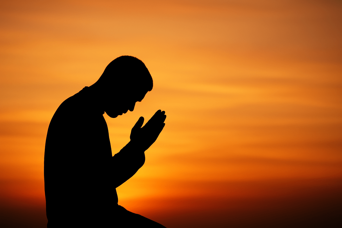 Silhouette of a person praying against a vibrant orange sunset sky. The individual appears to be kneeling or sitting, with their head bowed and hands pressed together in front of their face, creating a peaceful and contemplative mood. The background is a gradient of orange and yellow hues, suggesting either sunrise or sunset.