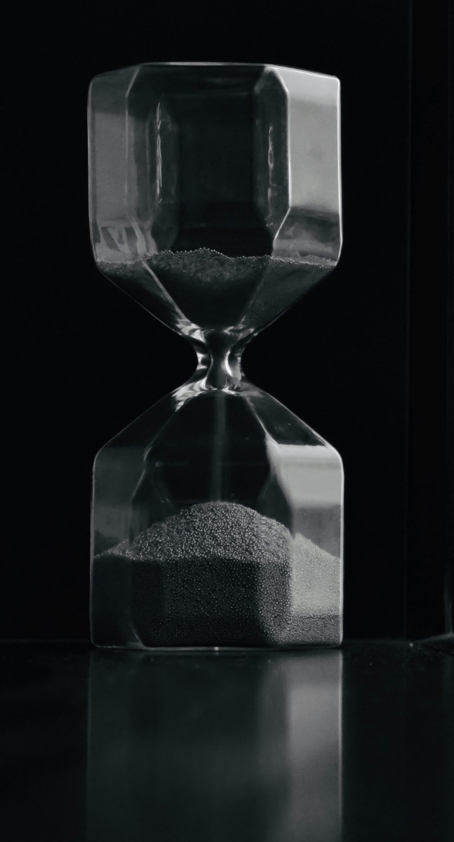 A clear glass hourglass filled with fine sand, set against a dark background. The hourglass has a geometric, faceted shape rather than the traditional rounded style. Most of the sand has flowed into the bottom chamber, with a small amount remaining in the top. The image is in black and white, and the lighting creates reflections and highlights on the glass surface, giving it a dramatic and elegant appearance. The hourglass is placed on a shiny, reflective surface.
